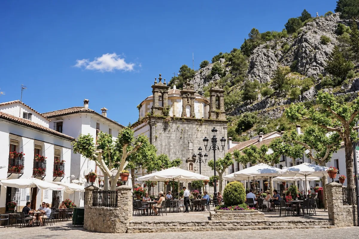 Terrazas con sombrillas blancas en la Plaza de España de Grazalema, frente a la fachada de piedra de la Iglesia de Nuestra Señora de la Aurora, con una ladera rocosa detrás
