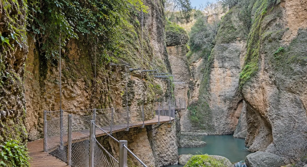 Pasarela de madera con barandilla metálica anclada a la pared de roca del Tajo, sobre el agua verde del río Guadalevín