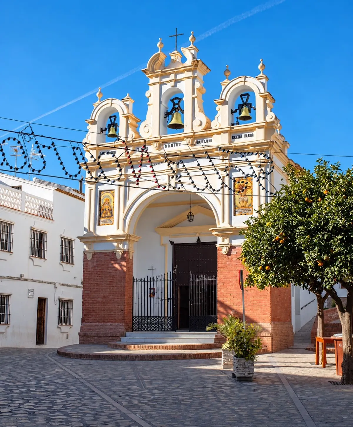 La Capilla de San Juan de Letrán, una capilla blanca con espadaña de tres campanas y zócalo de ladrillo, en una plaza empedrada del centro de Zahara de la Sierra, junto a un naranjo